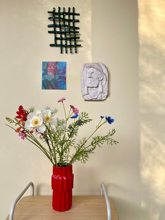 Vaaleankeltainen seinä, seinällä taidetta. Edessä pöytä, jonka päällä punainen maljakko ja niittykukkia.