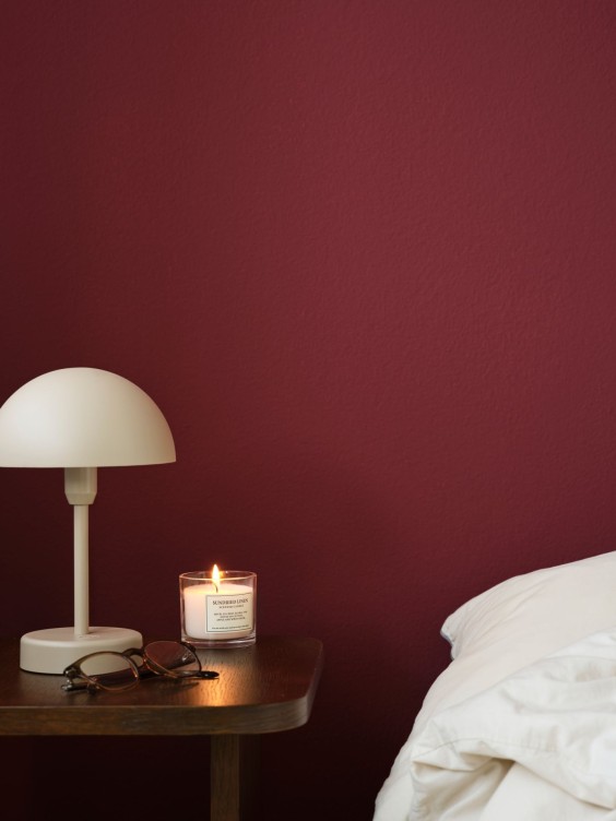 Elysia seinällä. Sängyn kulma ja yöpöytä, jossa valaisin ja kynttilä.