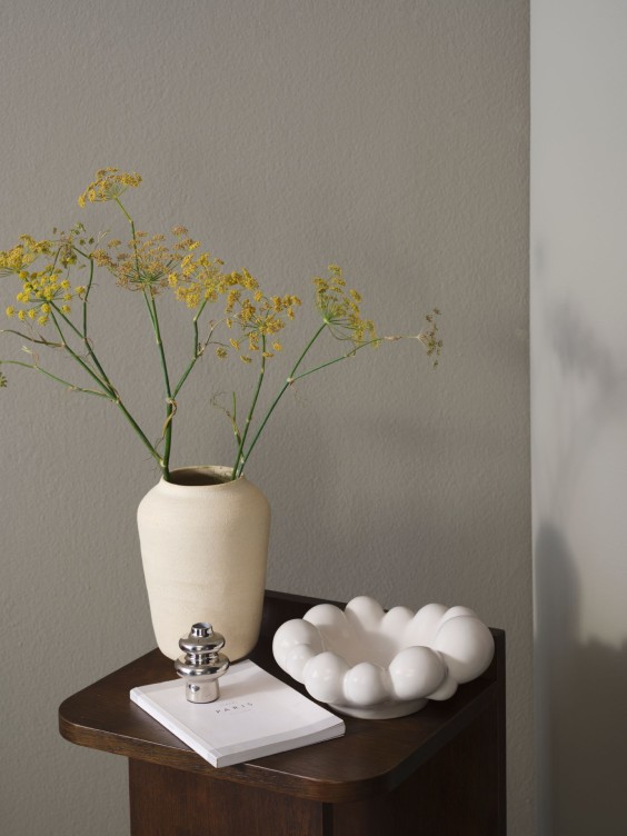 Mute Serenvale seinällä, edessä pieni pöytä, jossa vaalea maljakko.