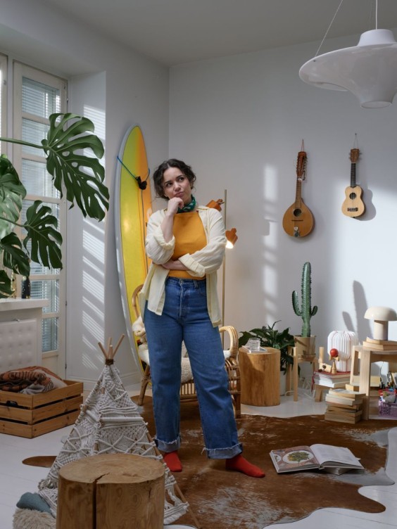 Olohuone ennen muodonmuutosta. Vaalea huone, jossa on paljon pientä tavaraa seinillä ja lattialla. Nainen seisoo huoneen keskellä mietteliäänä.