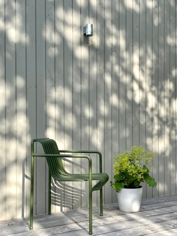 Vaaleanharmaa talon puuseinä ja terassi. Terassilla vihreä, metallinen tuoli ja kasvi.