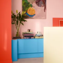 Oranssinpunainen seinä, sininen lipasto ja keltainen pöytä. Seinällä värikäs maalaus.