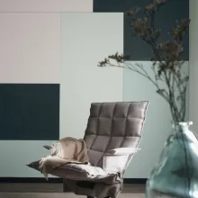 Mintunvihreitä, vaaleita ja tummia ruutuja seinällä. Edessä minimalistinen harmaa tuoli ja lasimaljakko.