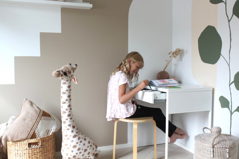 Tyttö istuu piirtämässä lastenhuoneen pöydän ääressä, kirahvi-pehmolelu