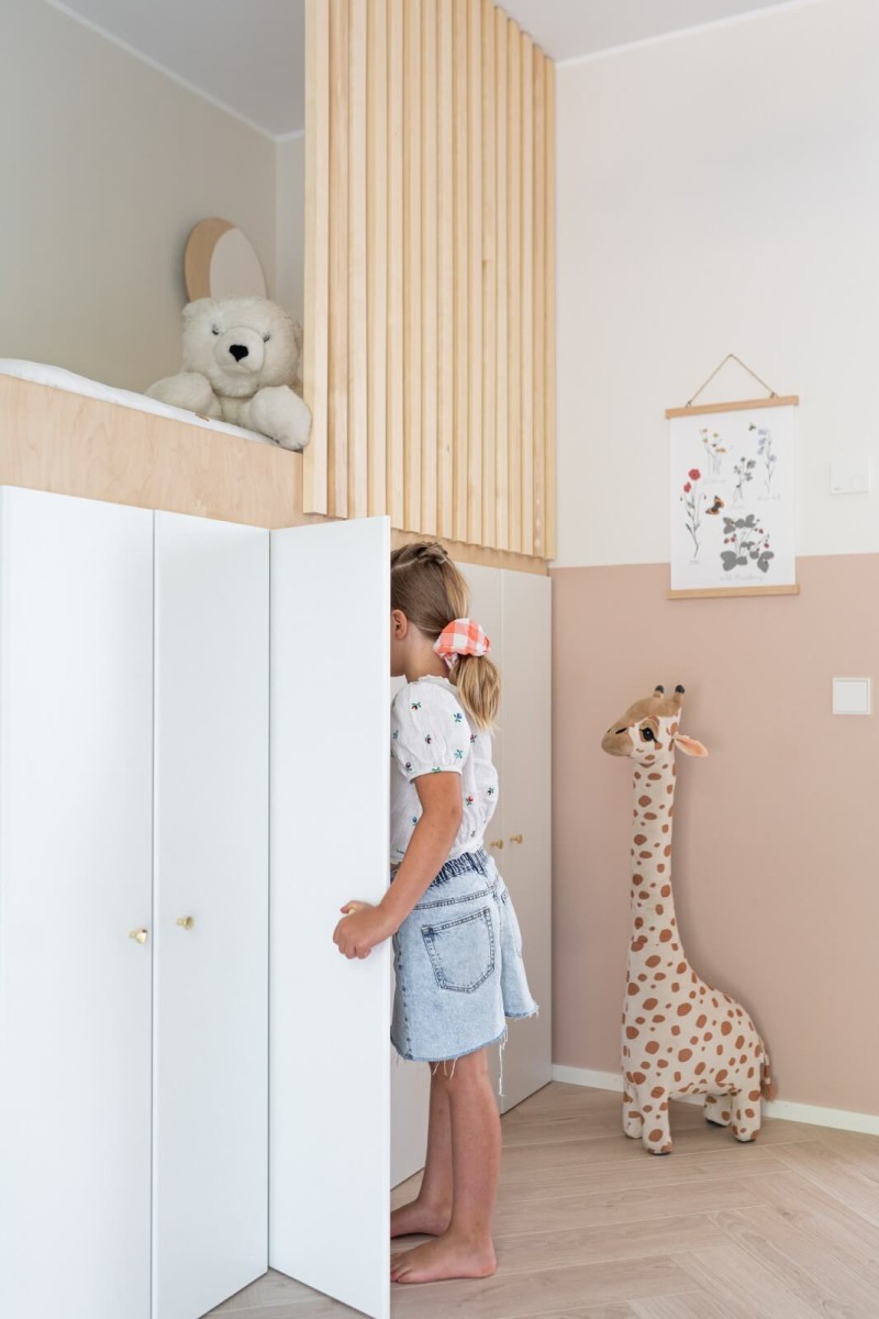 Tyttö kurkistaa vaatekaappiin lastenhuoneessa, moniväriset seinät, kirahvi-pehmolelu